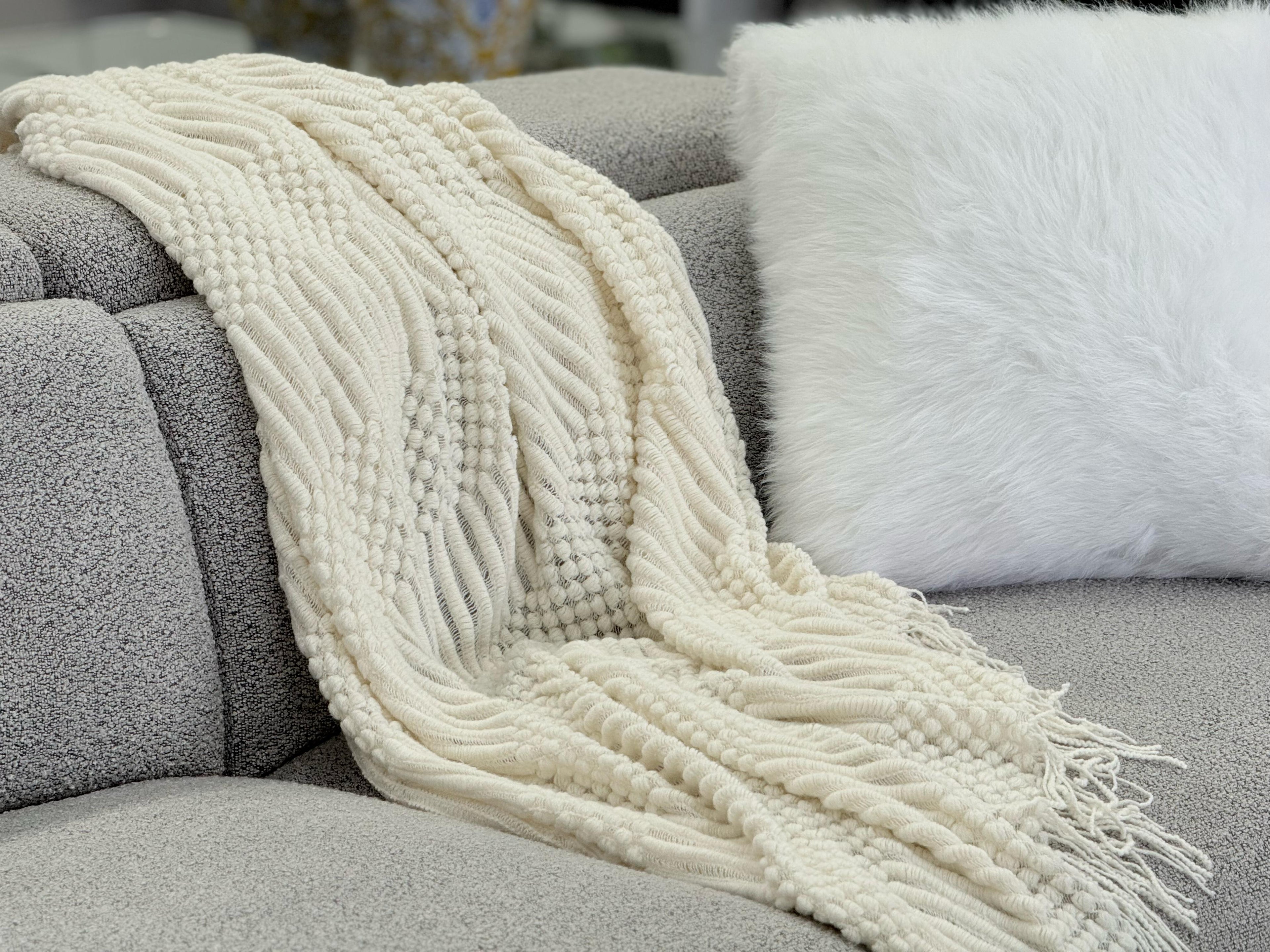 Cream Tessa throw rug blanket draped over a grey sofa with a fluffy white cushion for a cozy touch.