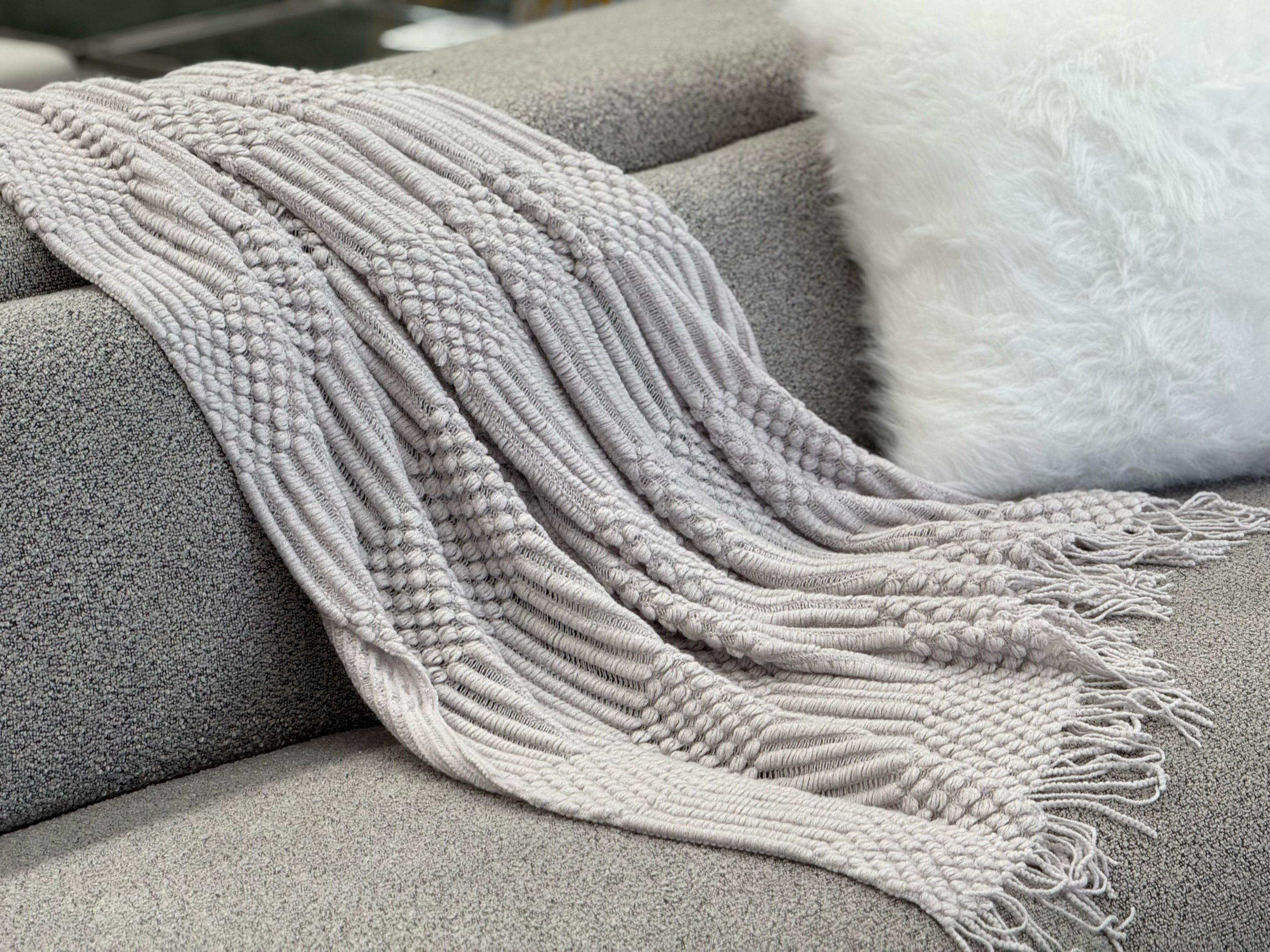 Grey Tessa throw rug blanket laid over a blue sofa, featuring a detailed textured weave and fringe ends.