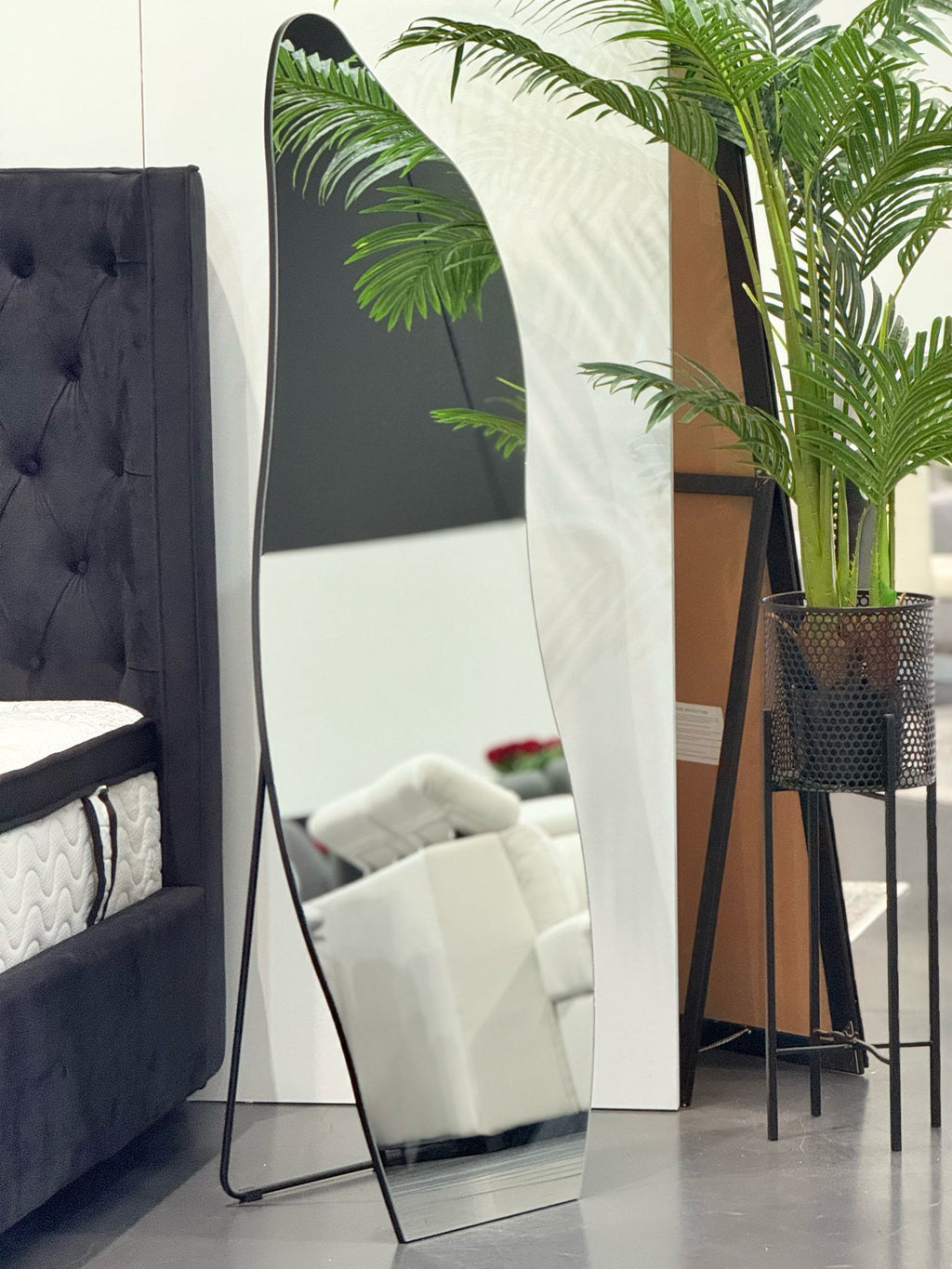 Irregular free standing mirror styled in showroom beside bed and indoor plant décor arrangement.