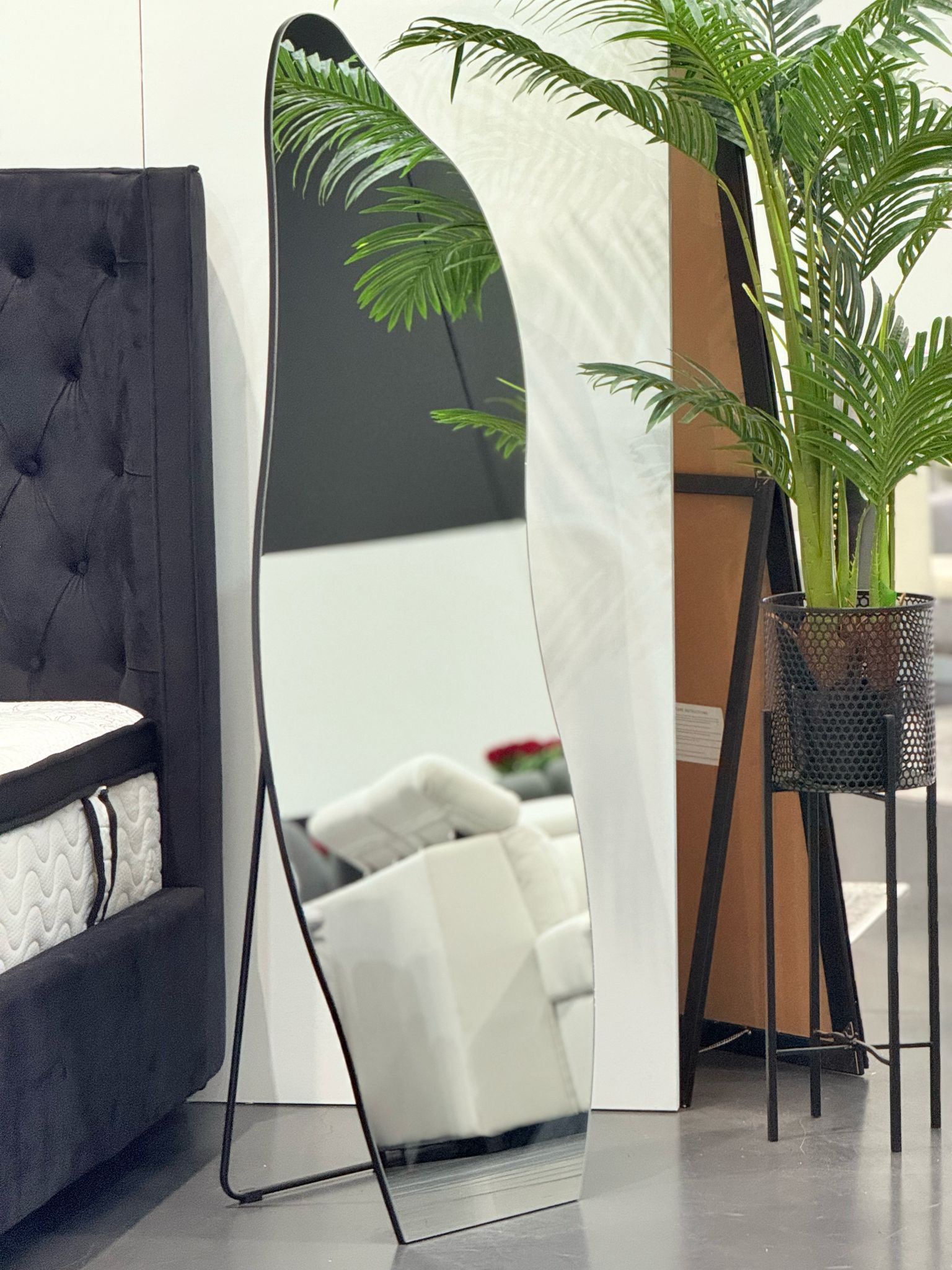 Irregular free standing mirror styled in showroom beside bed and indoor plant décor arrangement.